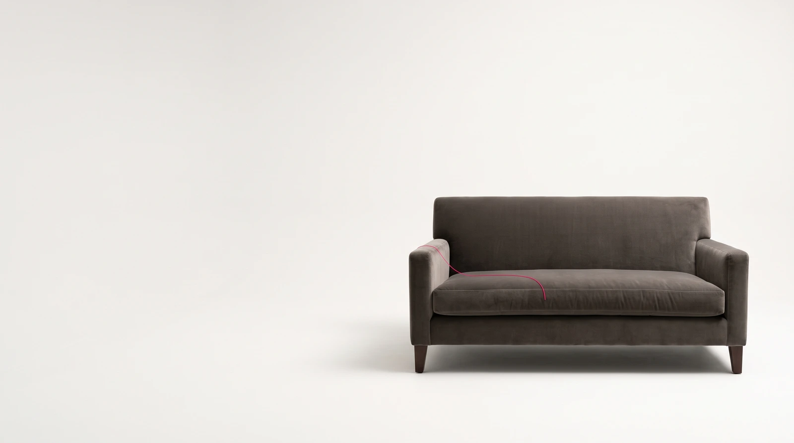 One low warm-grey velvet sofa standing alone on an empty pale studio sweep, photographed straight on, with a single fine magenta thread trailing from its near arm across the seat cushion and nothing else in the room